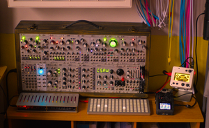 A warm, sharp picture shot from a slight downward angle, showing a big wooden
case full of silvery Eurorack modules knobs, dials, buttons and lights.
In front of the case are two silver devices, one with sixteen sliders and the
other with a grid of buttons, some of which are glowing.
Beside these are two small devices with screens, one of which is glowing a
bright amber color.
Cables in trans colors drape over the right-hand side.